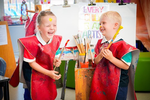 Laughing girl and smiling boy with paint on their face wearing smocks and holding paint brushes in front of easel