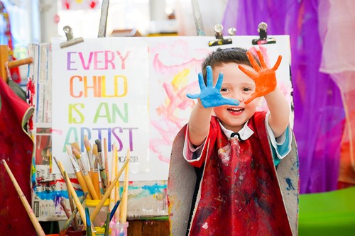 Young boy wearing a smock with blue and orange paint on his hands, standing in front of a painting